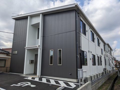 ダークグレーと白の外壁の２階建ての建物は敷地内には駐車場もあり、周りには民家がある。