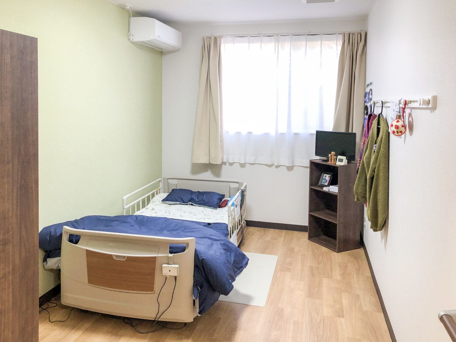 レースカーテンとカーテンが付いた窓があるフローリングの部屋には寝具付きのベッドや棚などが置かれている。