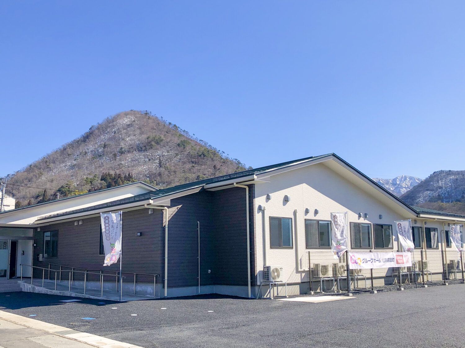 山が後ろにある平屋建ての建物の玄関先には手すり付きのスロープが付いている。