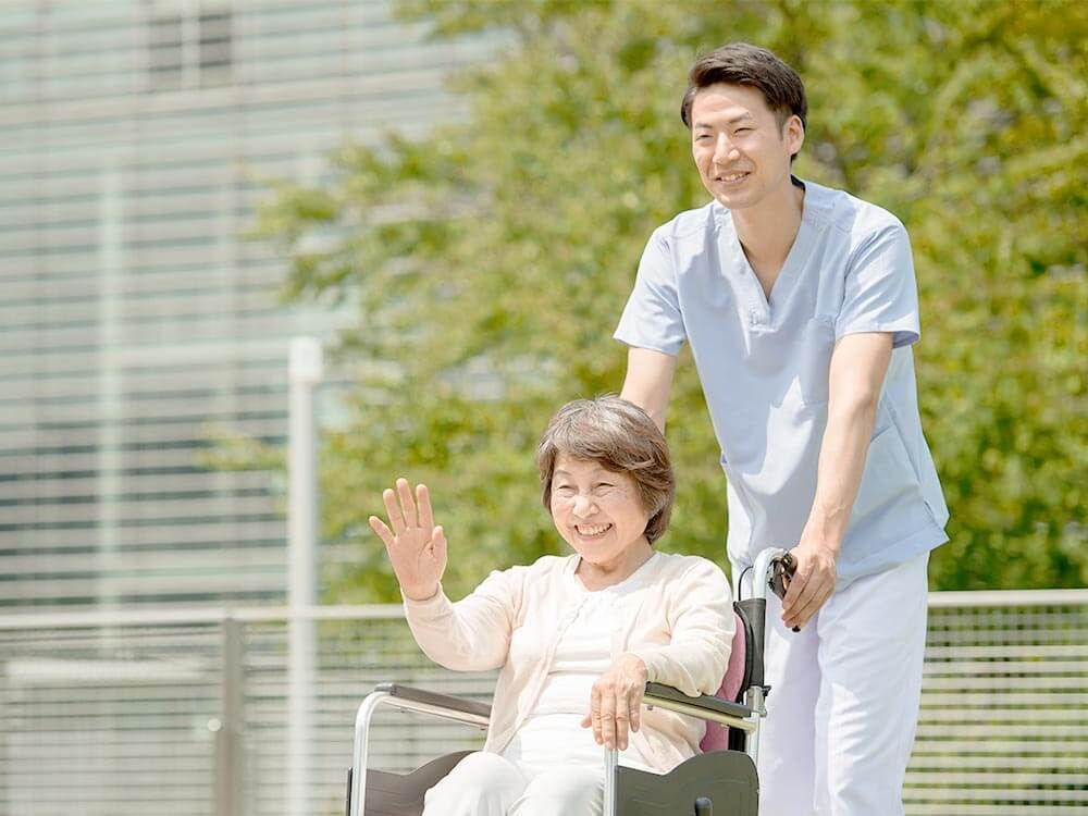 笑顔で誰かに手を振っている車いすの高齢女性と付き添いの男性スタッフ。日当たりのいい場所で。