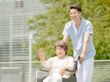 笑顔で誰かに手を振っている車いすの高齢女性と付き添いの男性スタッフ。日当たりのいい場所で。