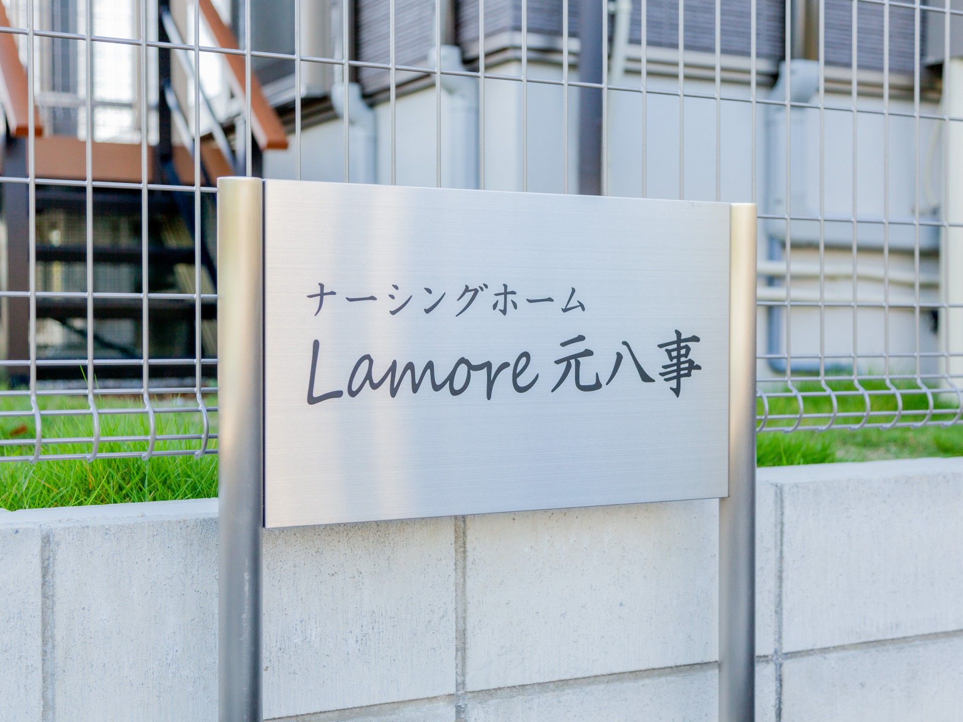 グレーの屋根が付いた２階建ての建物は敷地内に草木が植えられ、駐車場があり、玄関には屋根がある。