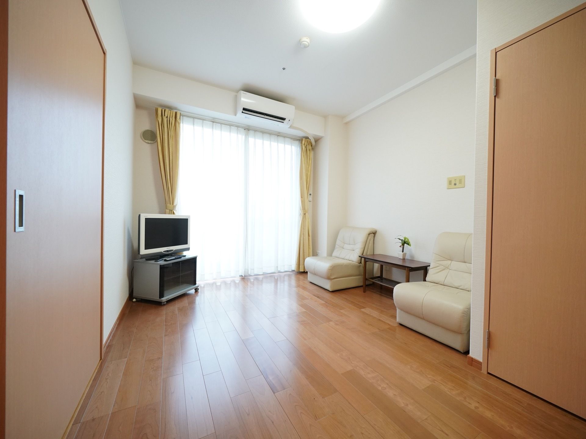 日当たりが良い居室で、部屋の隅にテレビが置かれている。大きな窓にはレースとカーテンが取りつけられている。