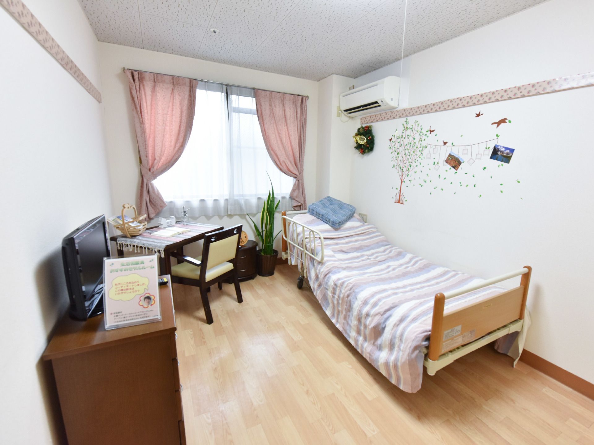 左側にはタンスの上にテレビが設置してあり奥にはテーブルと椅子もある。右手にはベッドがありエアコンもついている。