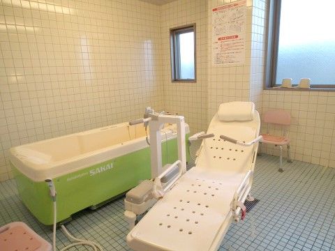 寝たまま入浴ができる特殊浴槽のリフト付き椅子と浴槽がある。浴室内はタイル張りであり、壁には機器の説明が書かれた看板がある。