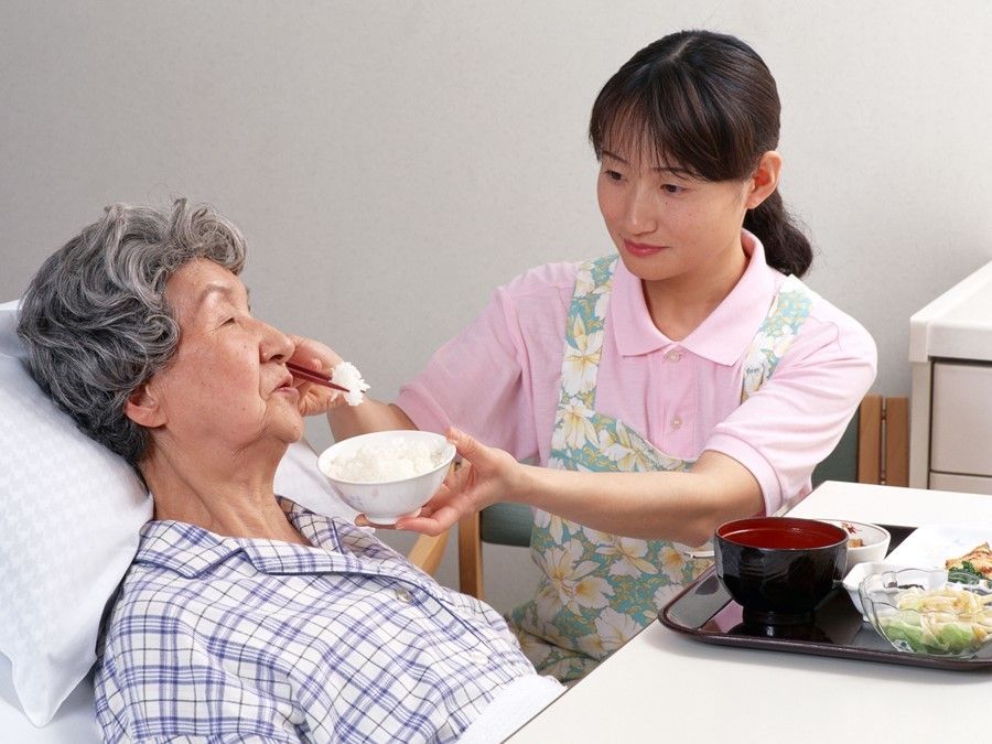 ベッドに座ったご高齢の女性が、介護スタッフにご飯を食べさせてもらっている様子。