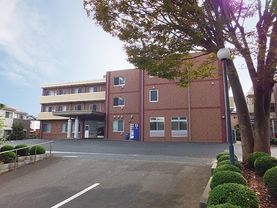 ３階建ての建物はオレンジ色の建物に白いバルコニーが付いた部分とバルコニーがない部分が繋がっている。玄関までは歩行者用通路が確保されている。