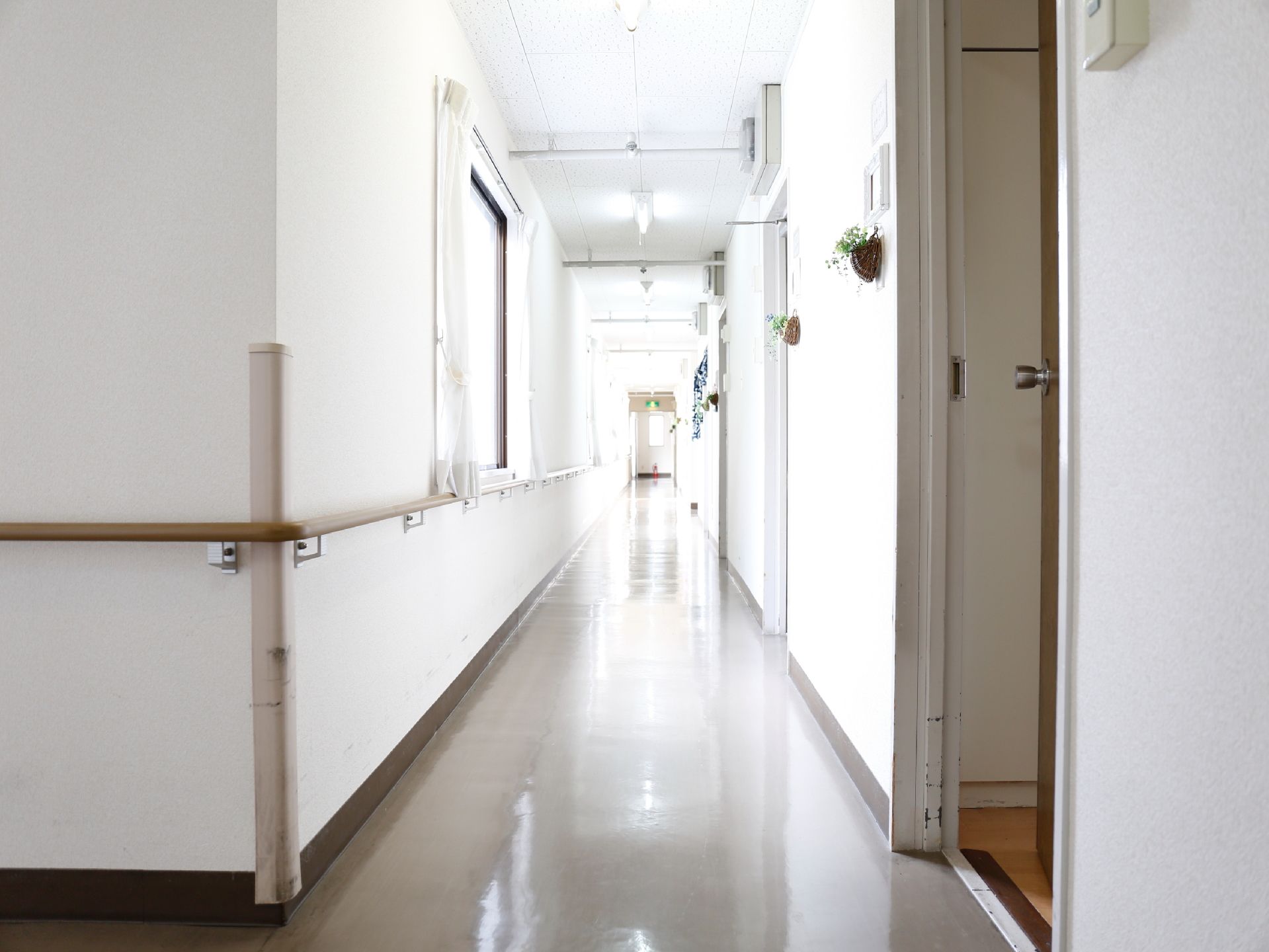 白い壁の廊下が長くのびており窓から光が差し込んでいる。手すりが左手に取り付けられており右手には扉がついている。