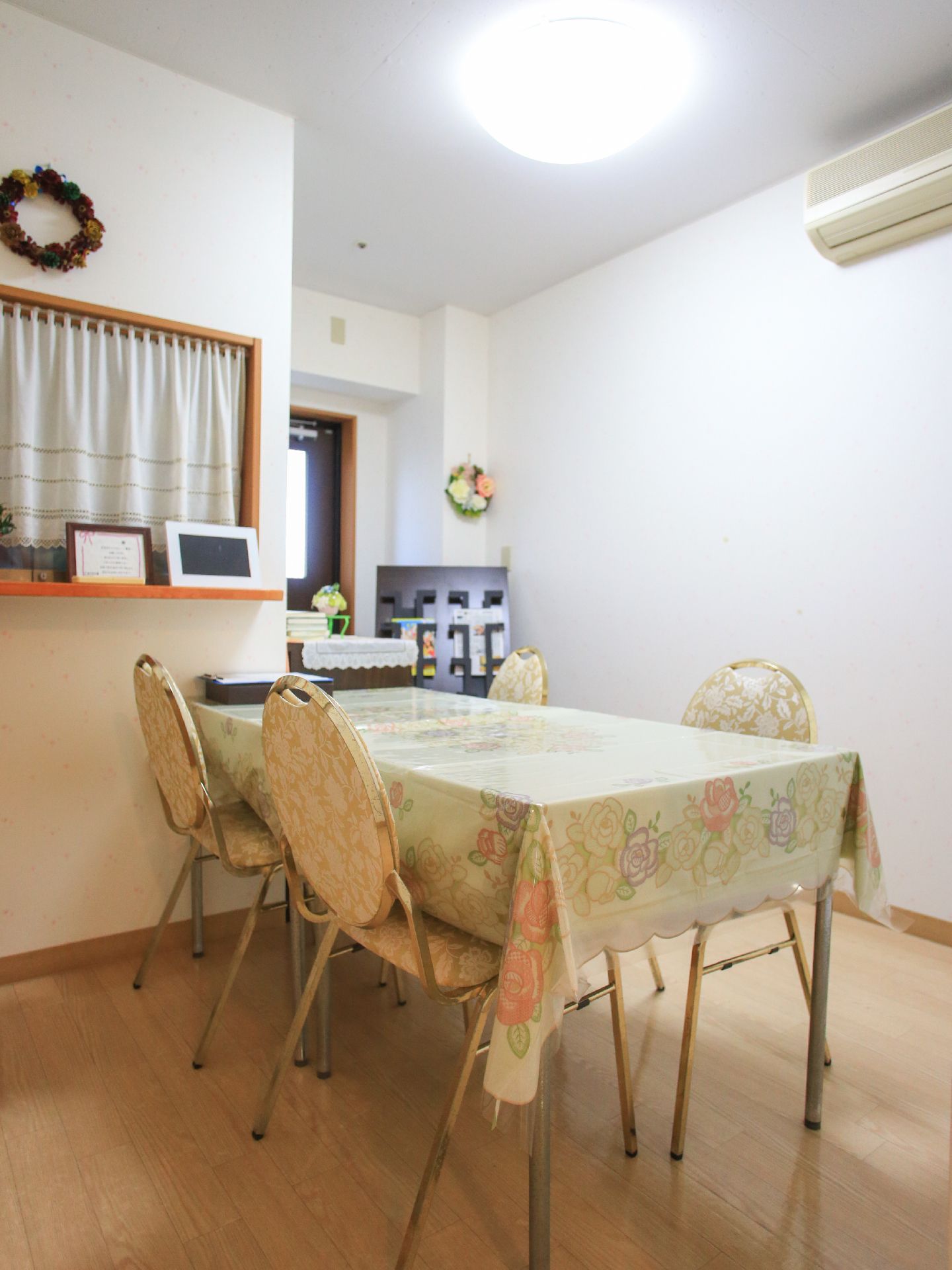 テーブルに花柄のクロスがかけられており、椅子が4脚用意されている。エアコンのついた部屋の床はフローリングになっている。