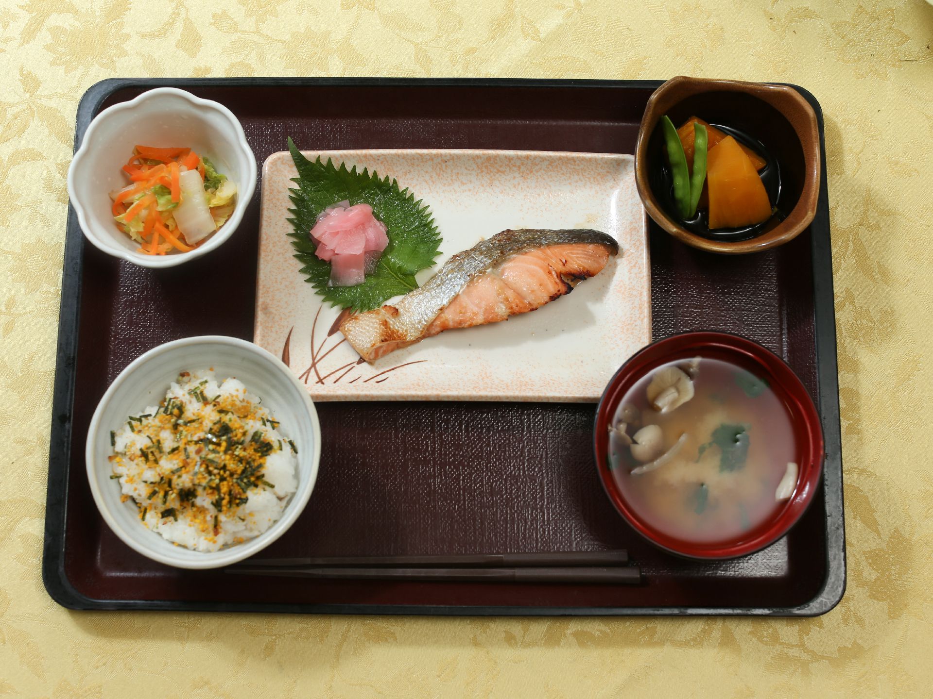 健康のバランスを考えた食事がトレイにのっている。シャケの切り身にかぼちゃの煮物などの小鉢が2品とごはんに味噌汁が置かれている。