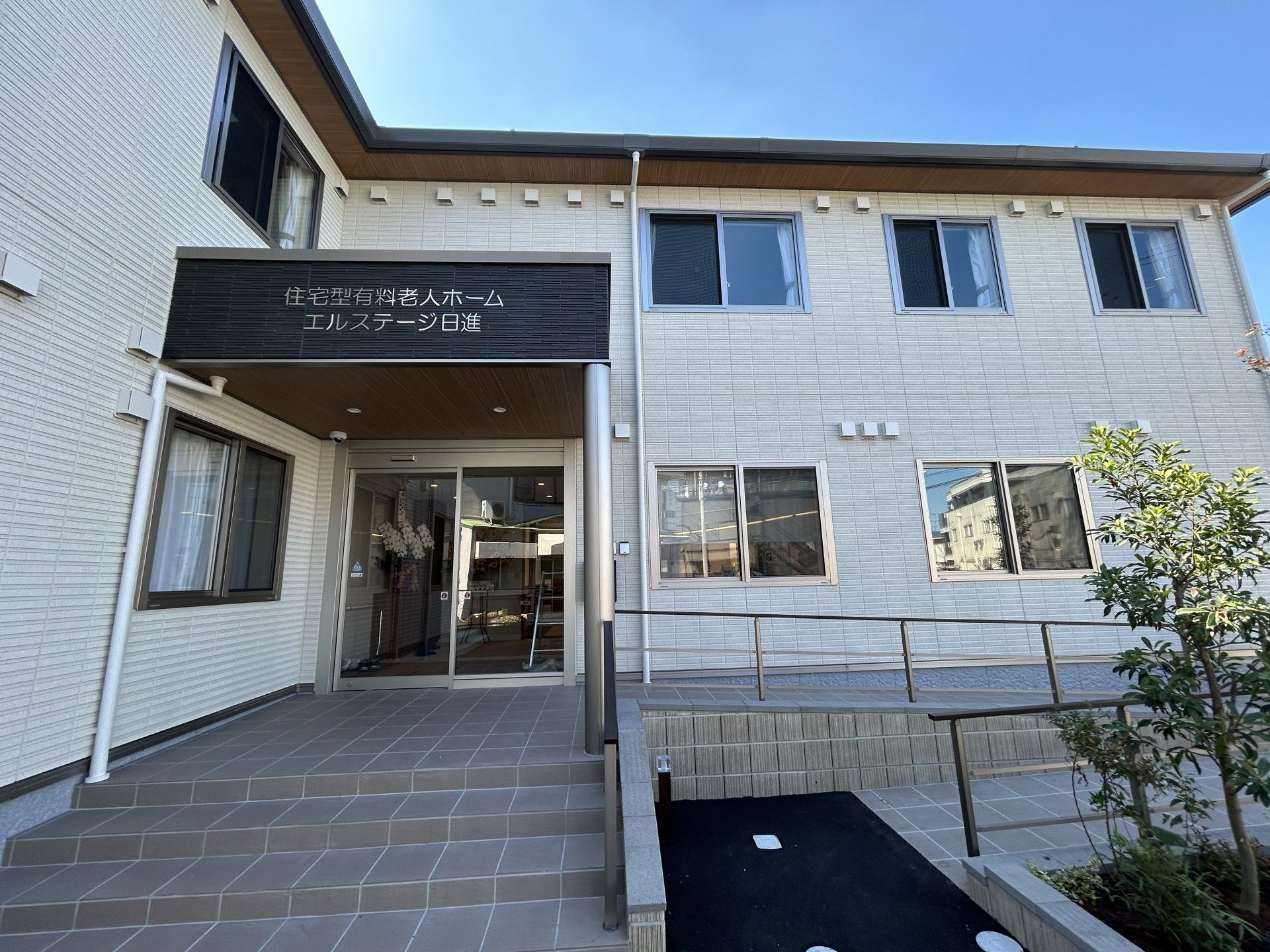 入り口に植物が植えられており、手すり付きの階段がある白い外壁の2階建て老人ホーム