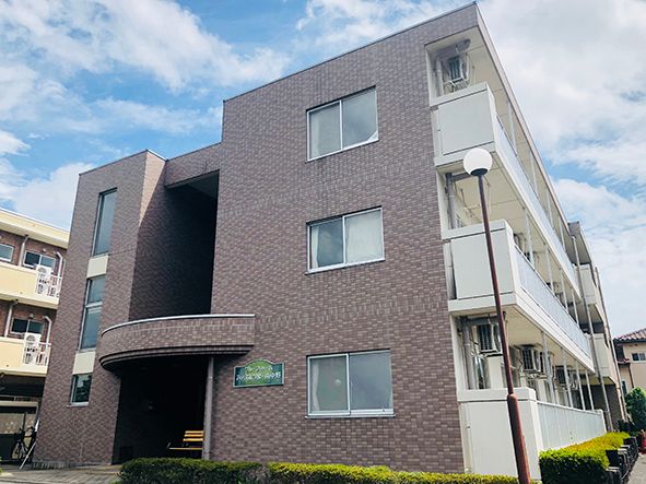 ３階建ての明るい茶色の外壁の建物で、建物廻りは舗装されて駐車場が整備されている。点字ブロックが備わっている。