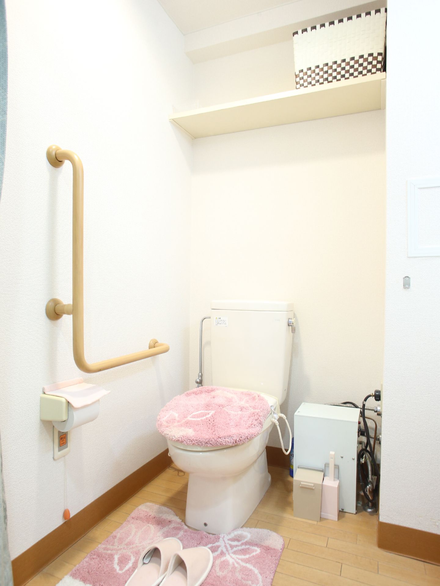 左手の壁にL字型の手すりとトイレットペーパーに呼び出しベルがついている。便座カバーのかかったトイレの上部には棚もついている。