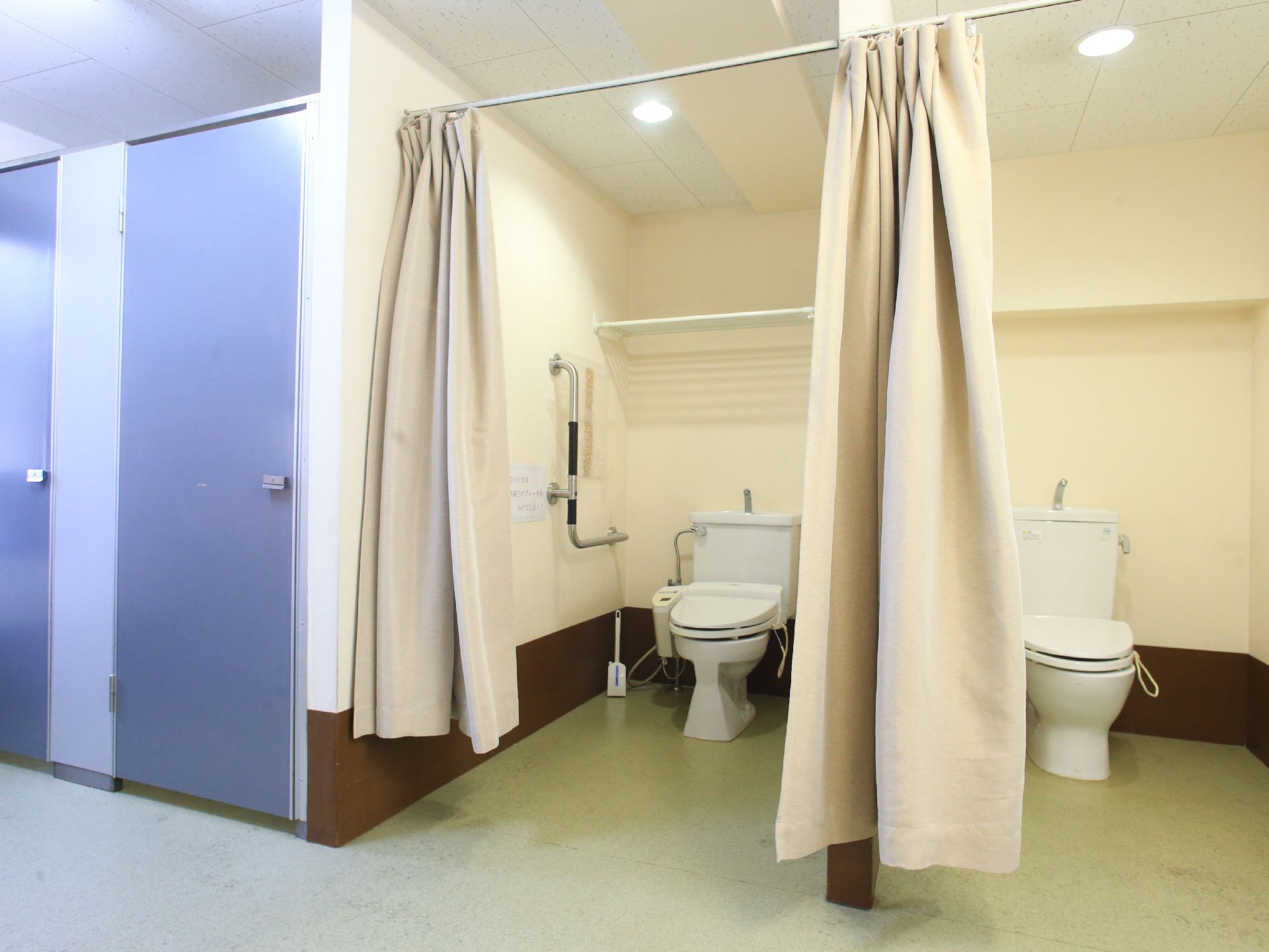 ウオッシュレットトイレが設置してあり手すりも壁についている。カーテンが仕切りの広々とした個室と、その左手に扉の個室がある。