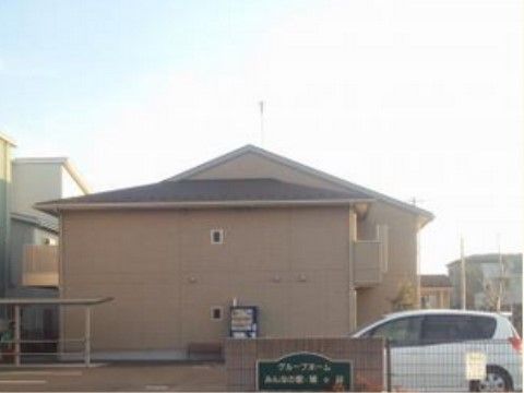 ベージュ系の２階建ての建物で、周囲の風景にも馴染んでいる。門には施設名が記されていて、広い駐車場がある。