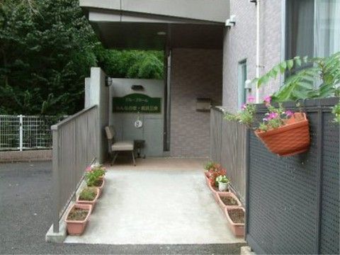 エントランスは緩やかなスロープがあり、手すりが付いている。入口前に長椅子が設置されている。プランターに植物が栽培されている。