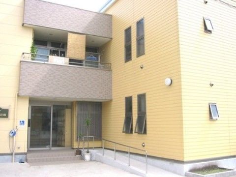 黄色の外壁に茶色のフェンスのエル型の２階建ての建物である。玄関前に２段の階段を設け、横側にスロープを設置している。