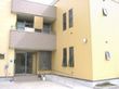 黄色の外壁に茶色のフェンスのエル型の２階建ての建物である。玄関前に２段の階段を設け、横側にスロープを設置している。