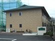 ブラウン系の２階建ての外観である。建物傍に自転車が１台あり、横側に緑の花壇を設けている。前方に表札を掲げている。