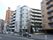 グレーの7階建ての建物になっておりベランダがみえる。道路に面して建っており目の前に樹木も植えられている。