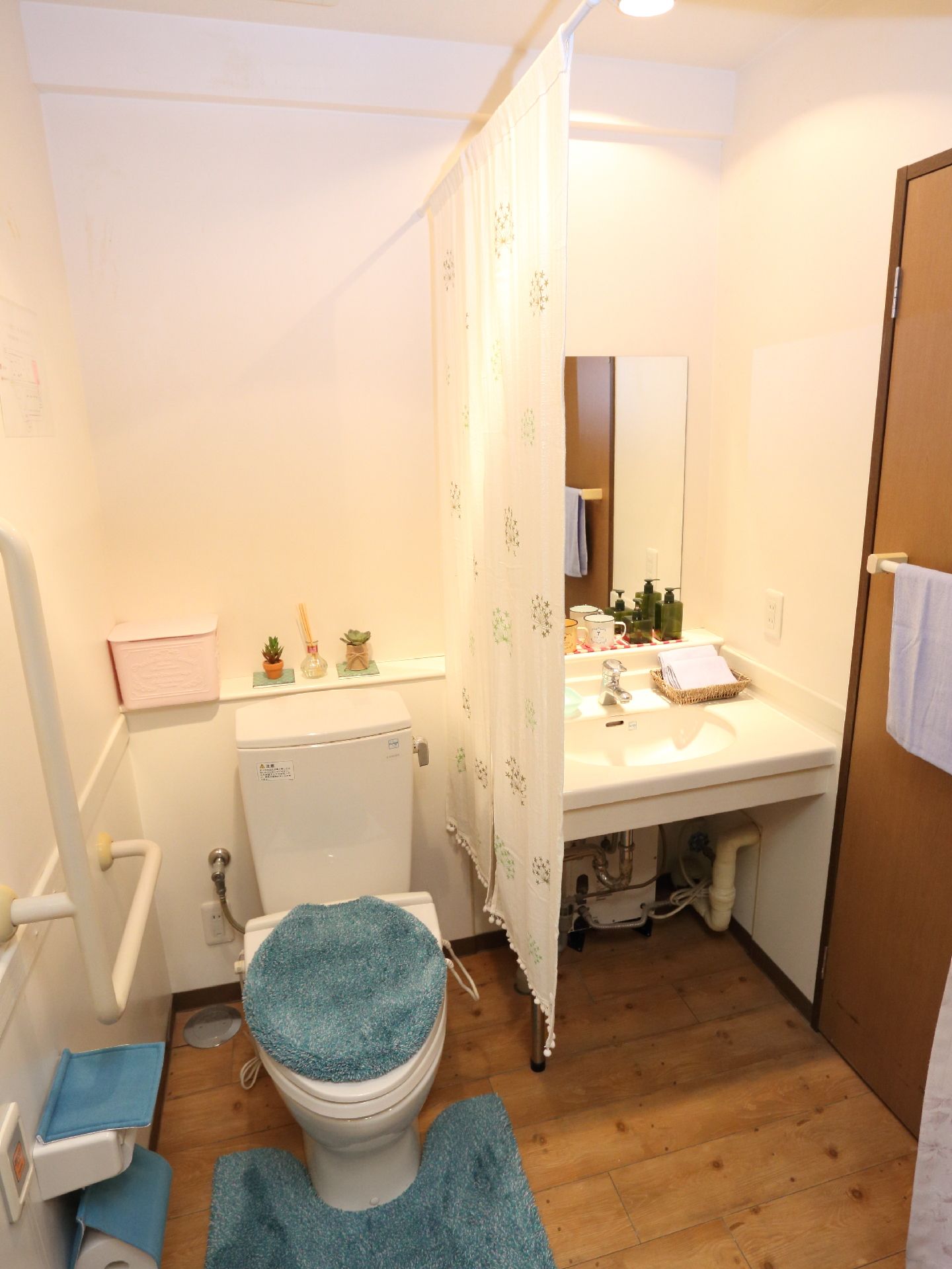 左側の壁にはL字型の手すりが設置してありトイレットペーパーフォルダーと便座にはブルーのカバーがしてある。右側は流しになっている。