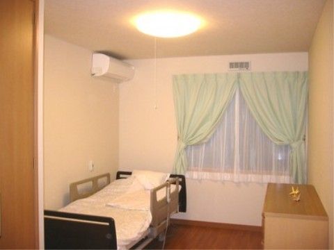 居室にはエアコン、介護用ベッドを備え、大きな窓には、薄いブルーのセンタークロスのカーテンを掛け、お洒落な雰囲気である。