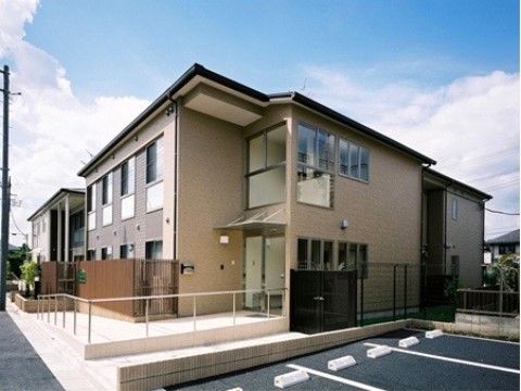 落ち着いた色合いのグループホームである。建物の横にはアスファルトで舗装された駐車場が設けられている。
