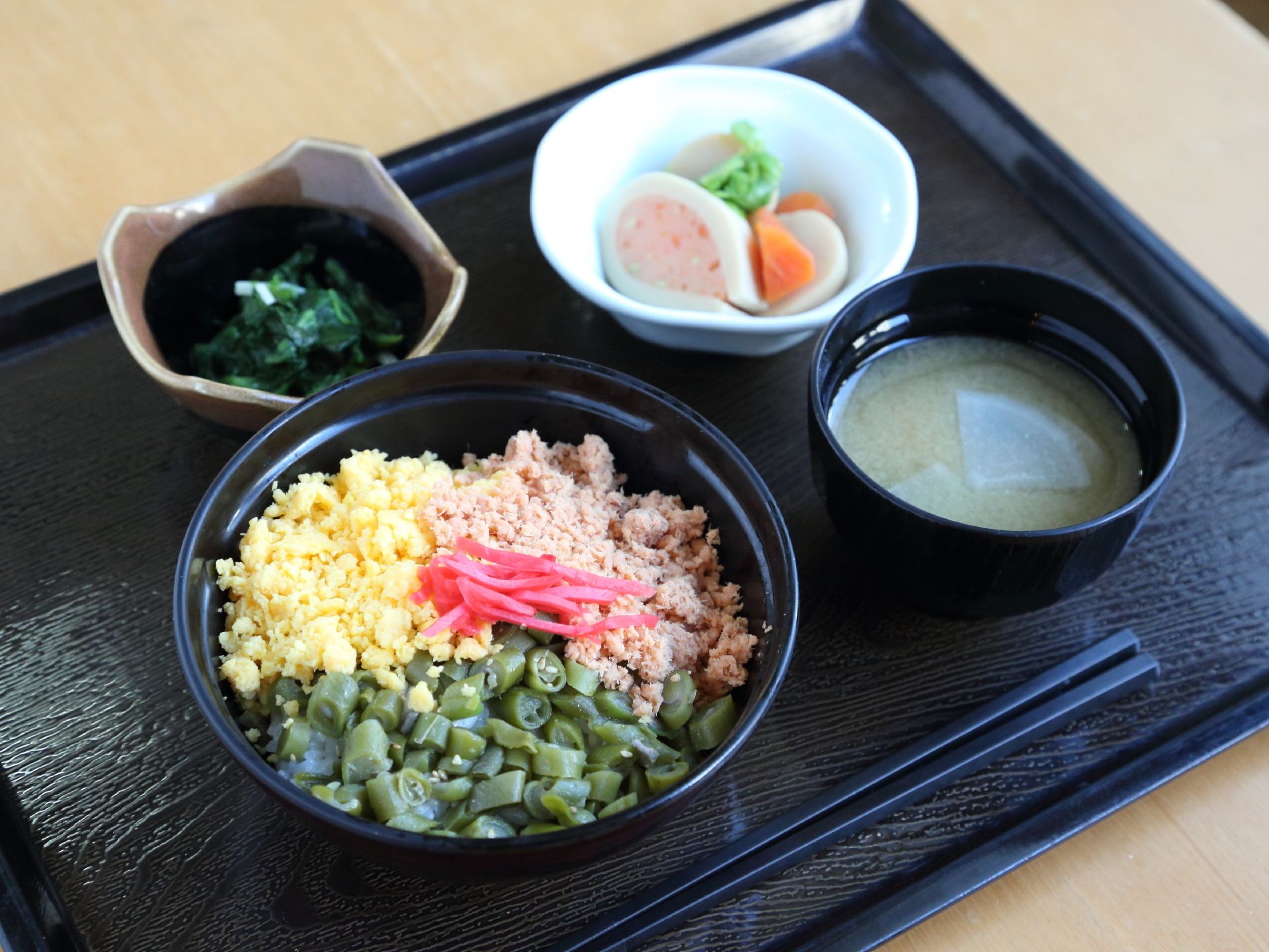栄養バランスを考えられた食事が黒いお盆にのって提供されている。3色の丼と2品の小鉢に味噌汁が用意されている。