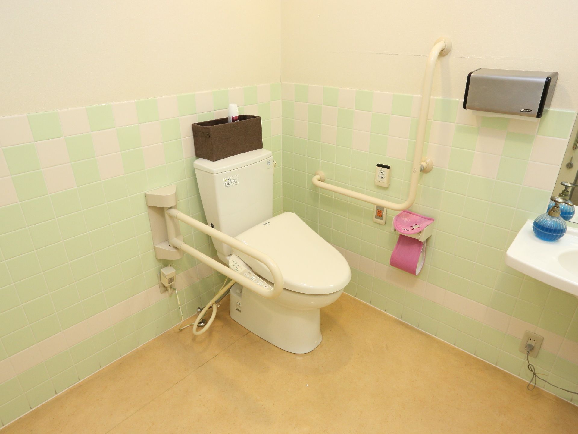 壁面は薄い緑と白でデザインされており右側にはトイレットペーパーとナースコールなども敷設されている。便器の反対側にも手すりがある。