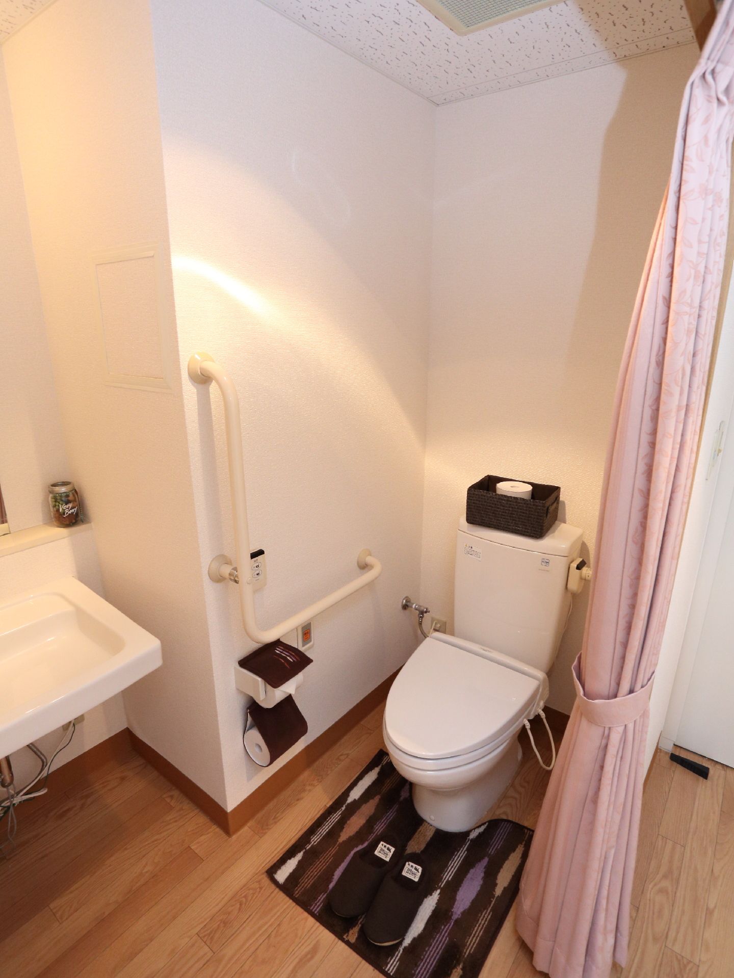 左手に流しがあり壁にはL字型の手すりとトイレットペーパーに呼び出しベルがある。タンクの上にカゴがあり扉はカーテン式になっている。