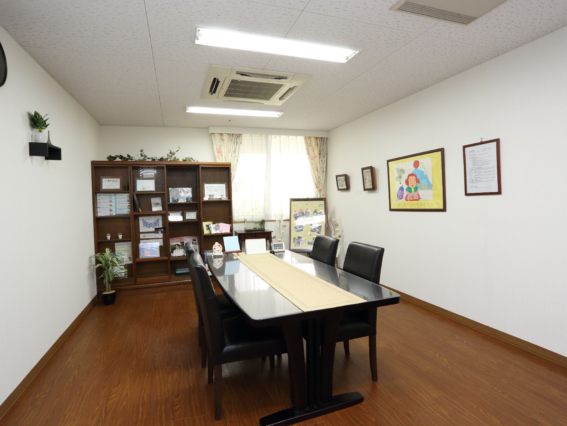 白と茶を基調とした面談室。中央にはテーブルと椅子、壁際には大きい棚が設けられている。窓と換気口も備わっている。