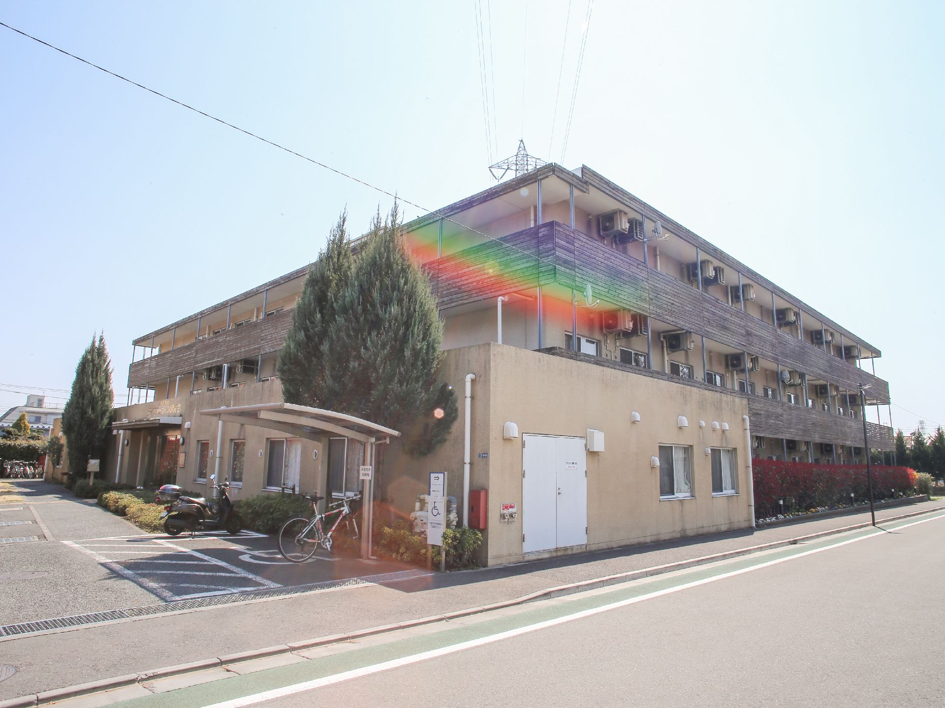 落ち着いたベージュ色の外観で、建物は3階建てになっている。駐輪場のあたりなどには、木々も植えられている。