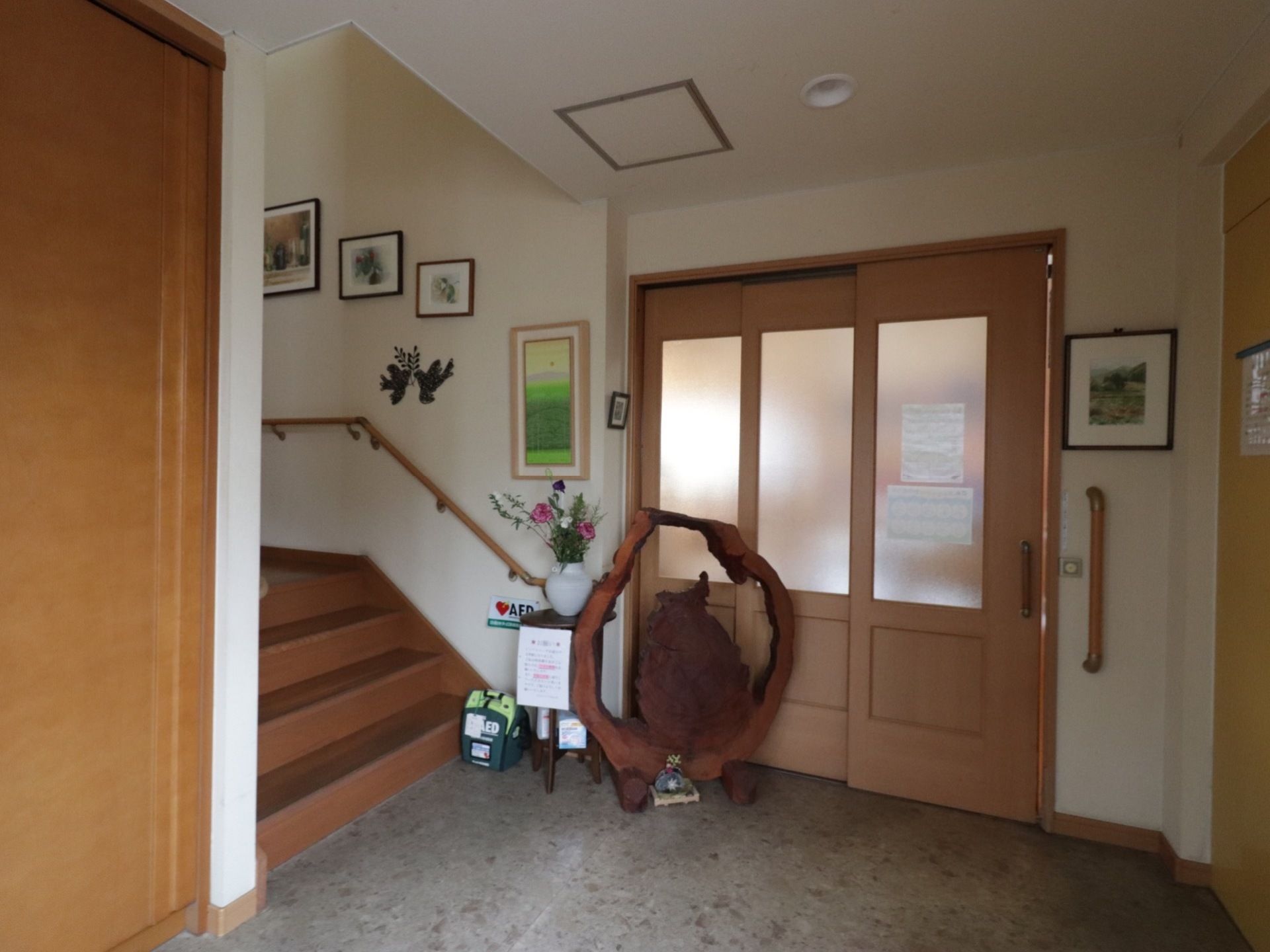 玄関ホールには、AEDが設置されている。木彫りの置物、花、絵画が飾られており、明るい雰囲気である。階段には手すりが設置されている。