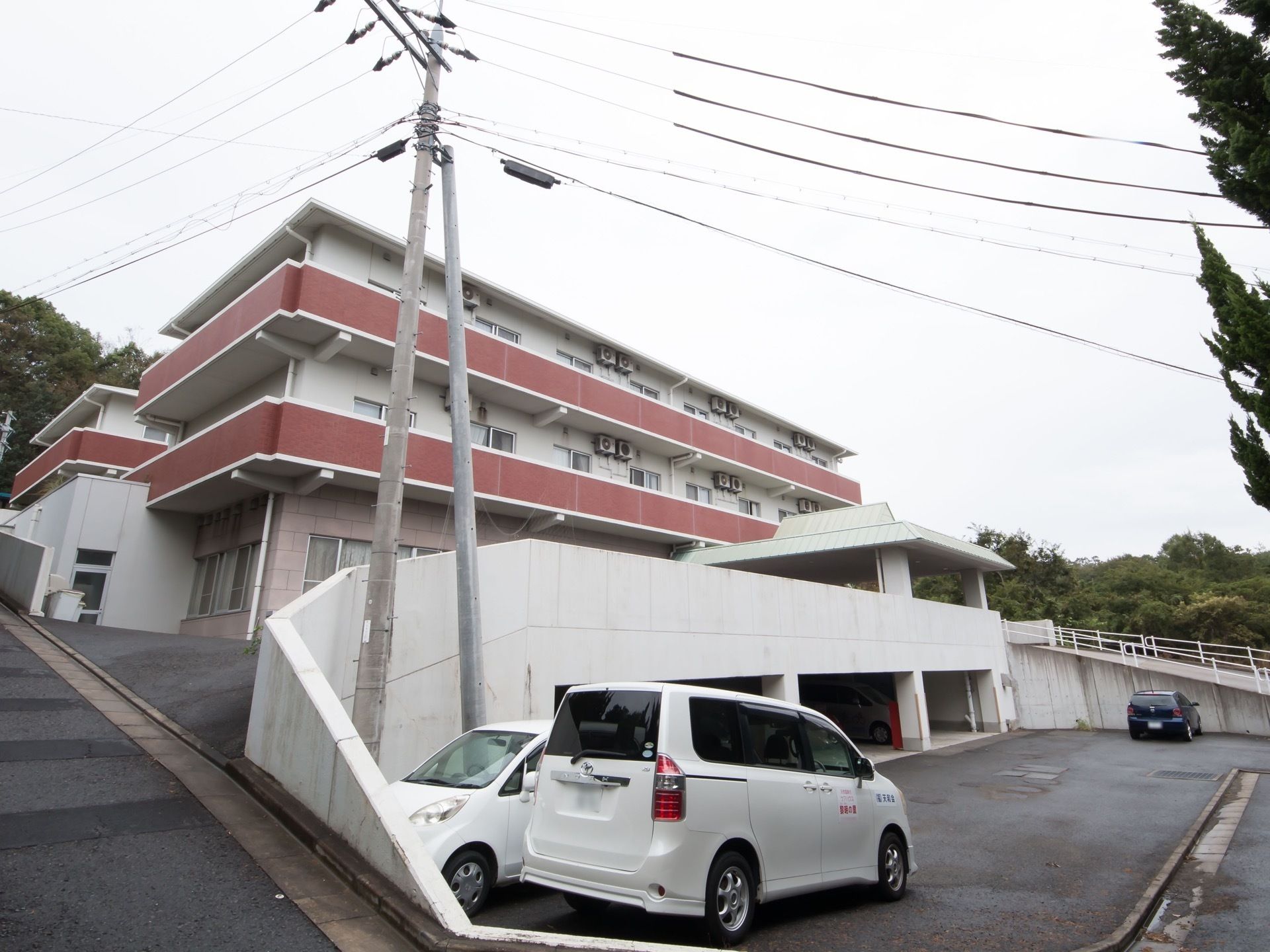 高台に白い外壁と、紅色のベランダ壁の建物が建っている。施設前には自動車が３台停められている。入口まで坂道がつながっている。