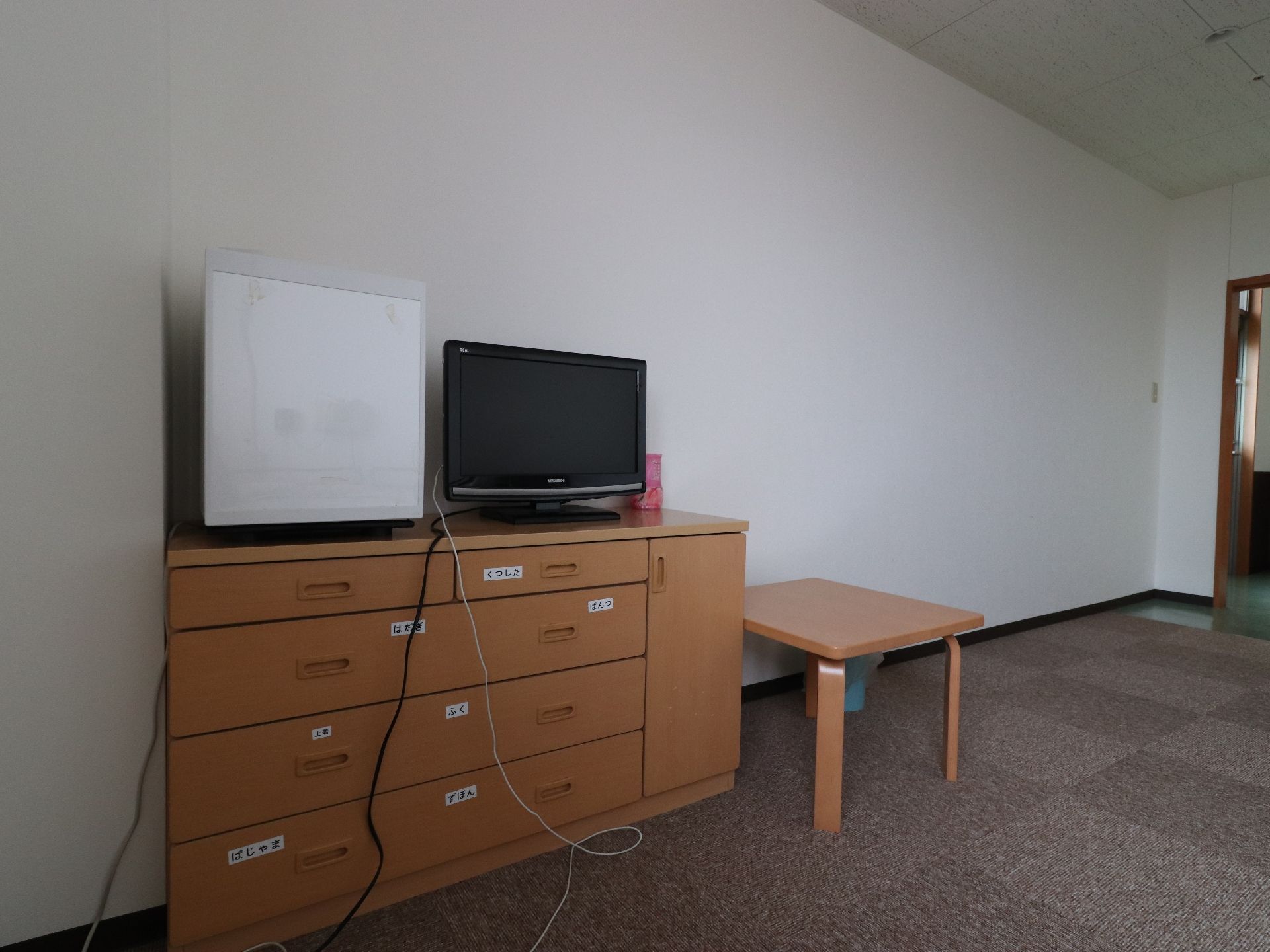 居室の中には白い壁を背にしてライトブラウンのチェストが置かれている。引き出しの前面には中に入れる物を書いたラベルがある。