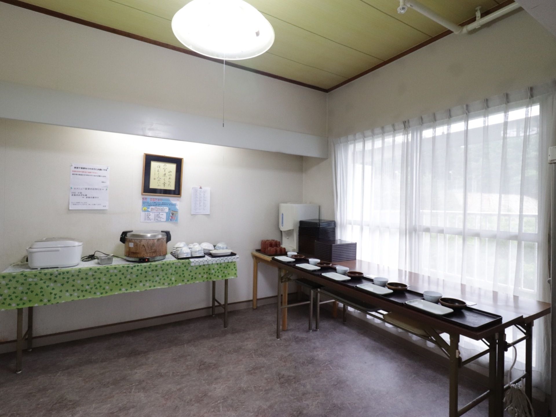 部屋のすみに、横長の折り畳みテーブルが設置されている。テーブルの上には、トレーに載った食器類や、炊飯器などがある。