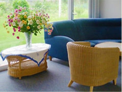 窓の傍にラタン製のサイドテーブルがあり、花が飾られている。壁際にソファとローテーブルを挟んでラタンの椅子が置かれている。