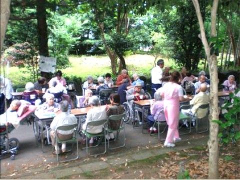 自然に囲まれた中に並べられたテーブルと椅子に座って食事を楽しむ入居者