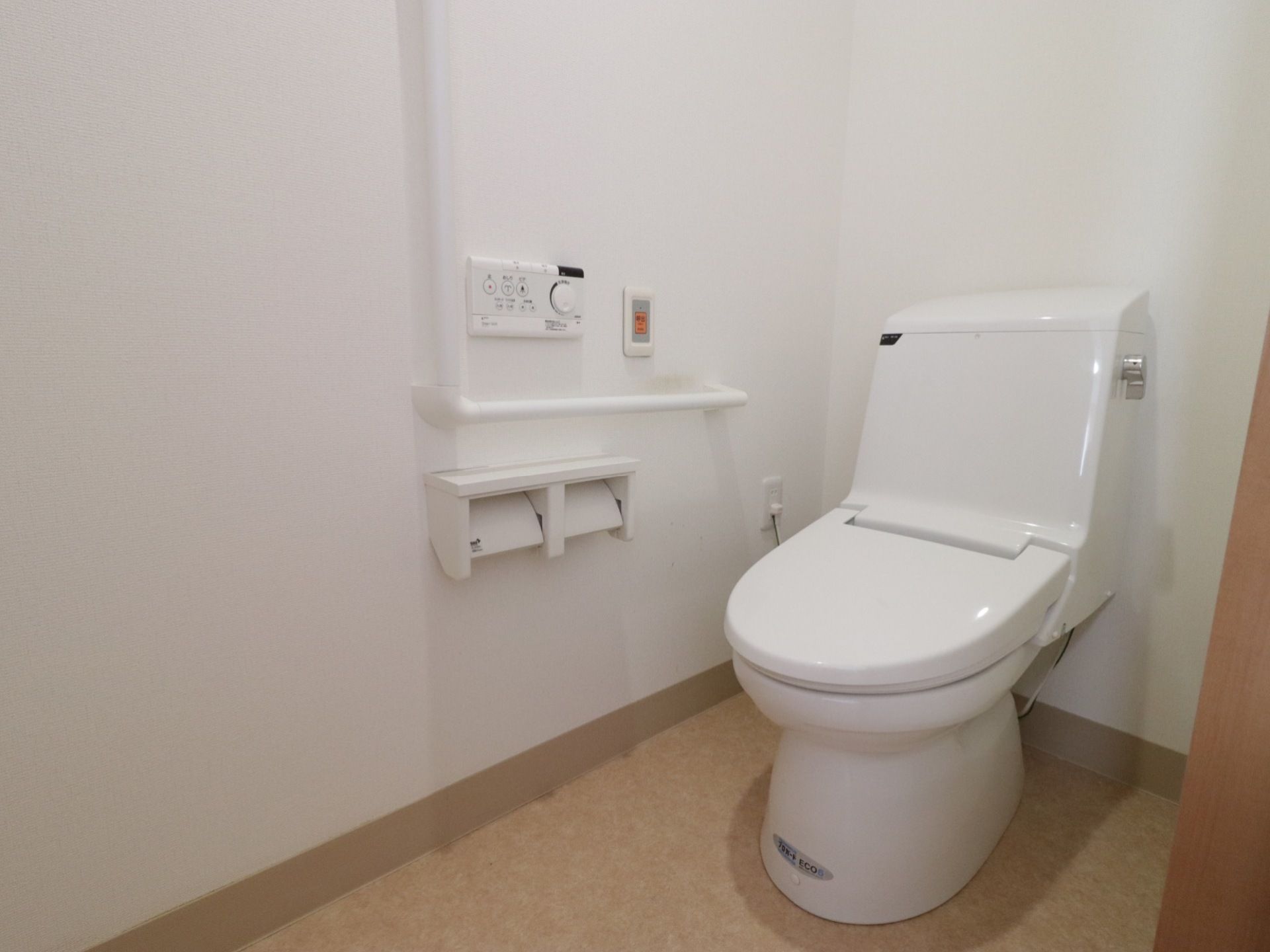 白を基調とした衛生的なトイレである。壁には手すり、非常ボタン、ペーパーホルダーと温水洗浄便座のリモコンが設置されている。