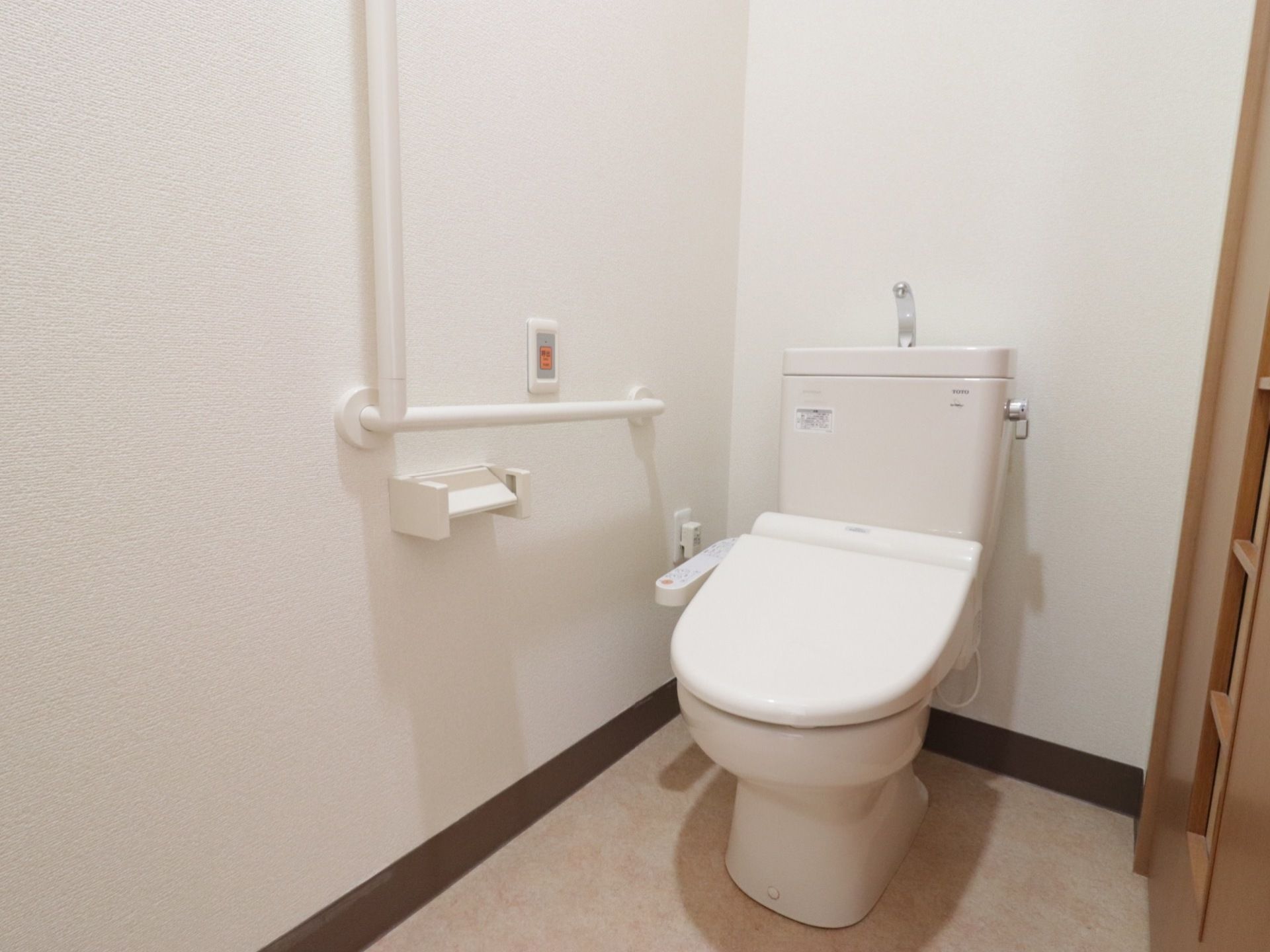 L字型の手すりが設置されたトイレである。白を基調としており、飾りがないシンプルなインテリアである。緊急時呼び出しボタンが備わっている。