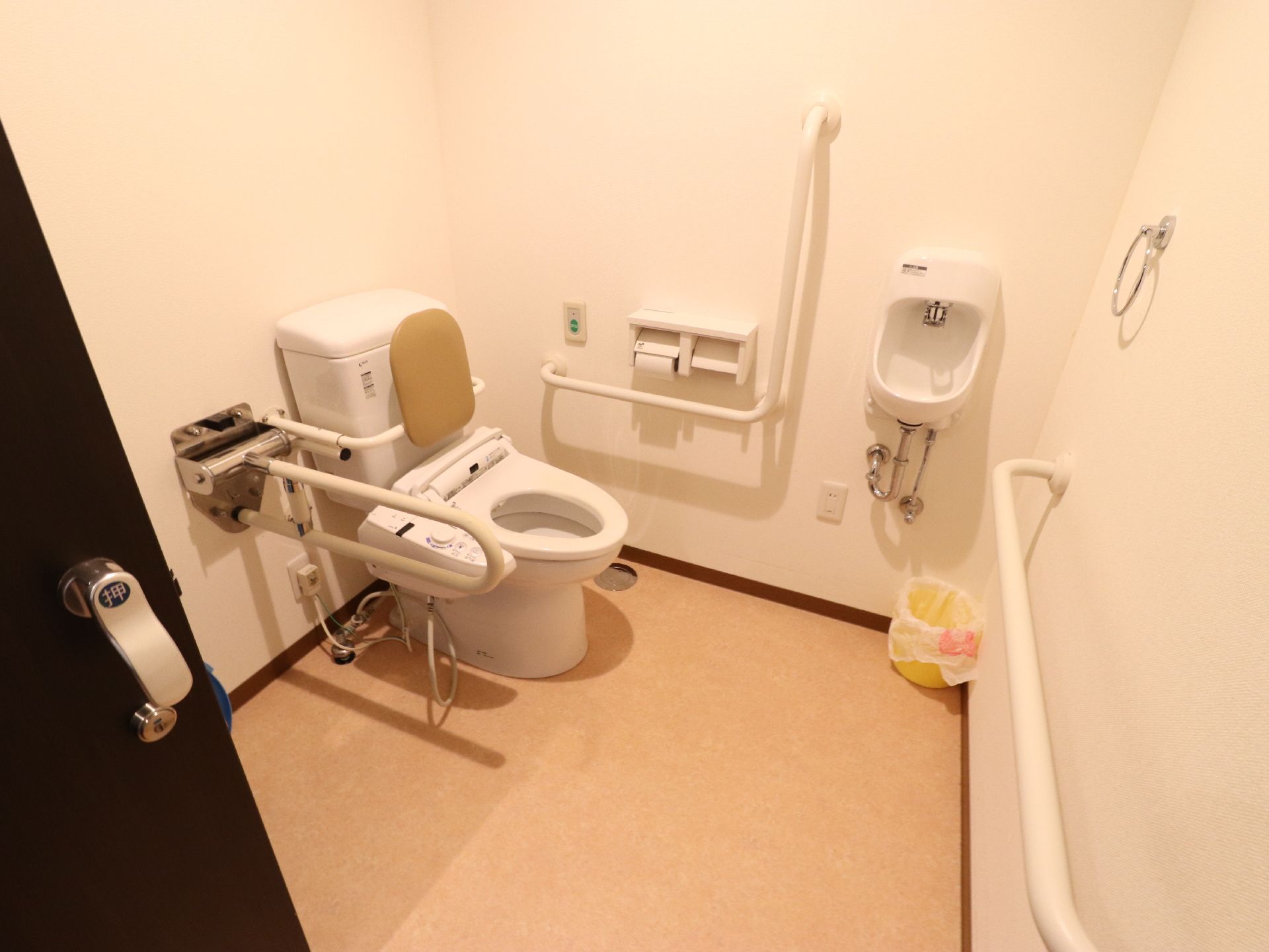 広々とした白い壁のトイレは綺麗で清潔感がある。手洗所、温水洗浄便座、緊急呼出ボタン、手摺が設置されている。
