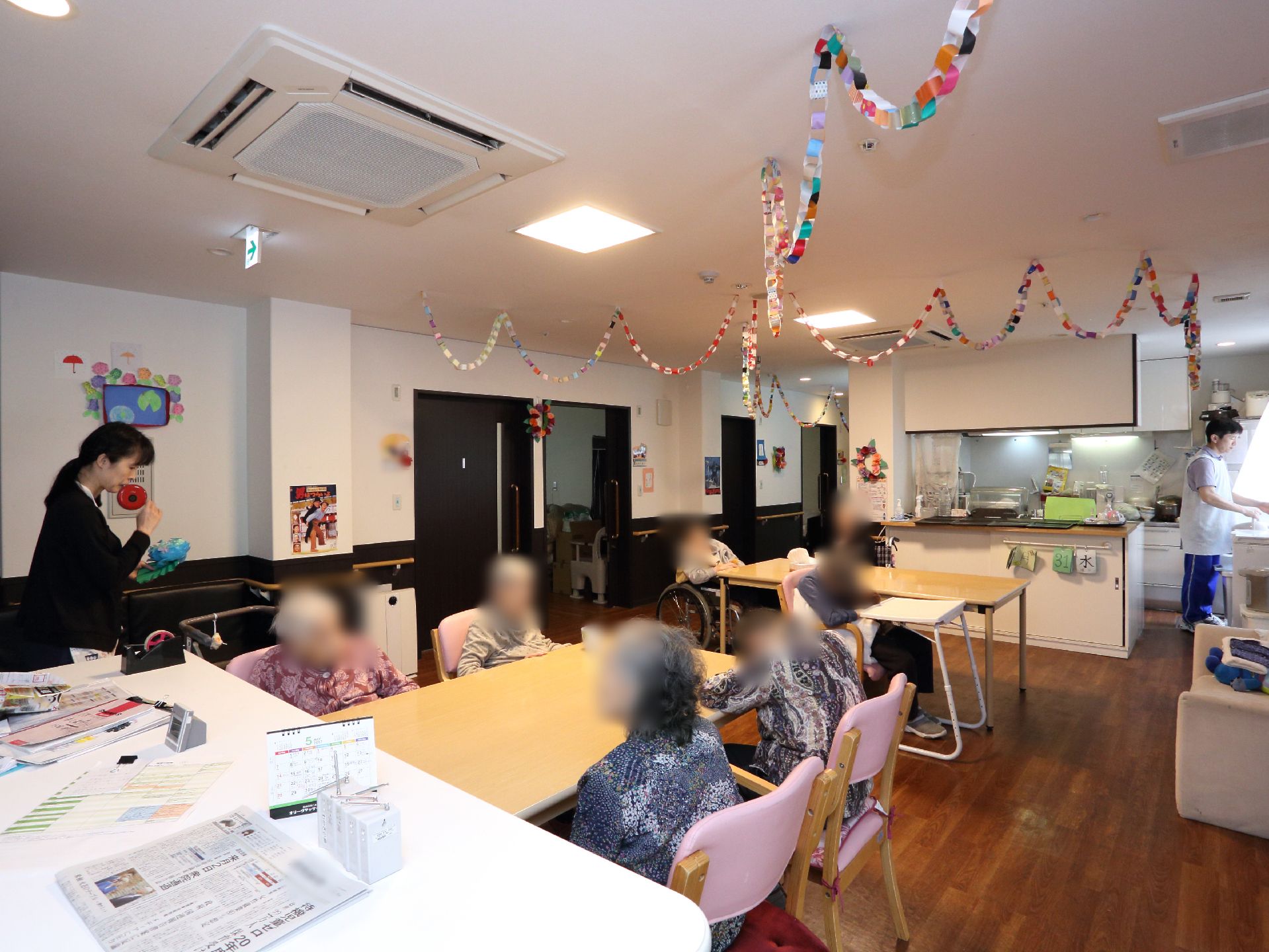 天井には空調が設置してあり手作りの装飾品が下がっている。テーブルのまわりを入居者やスタッフが囲んでおり談笑している。
