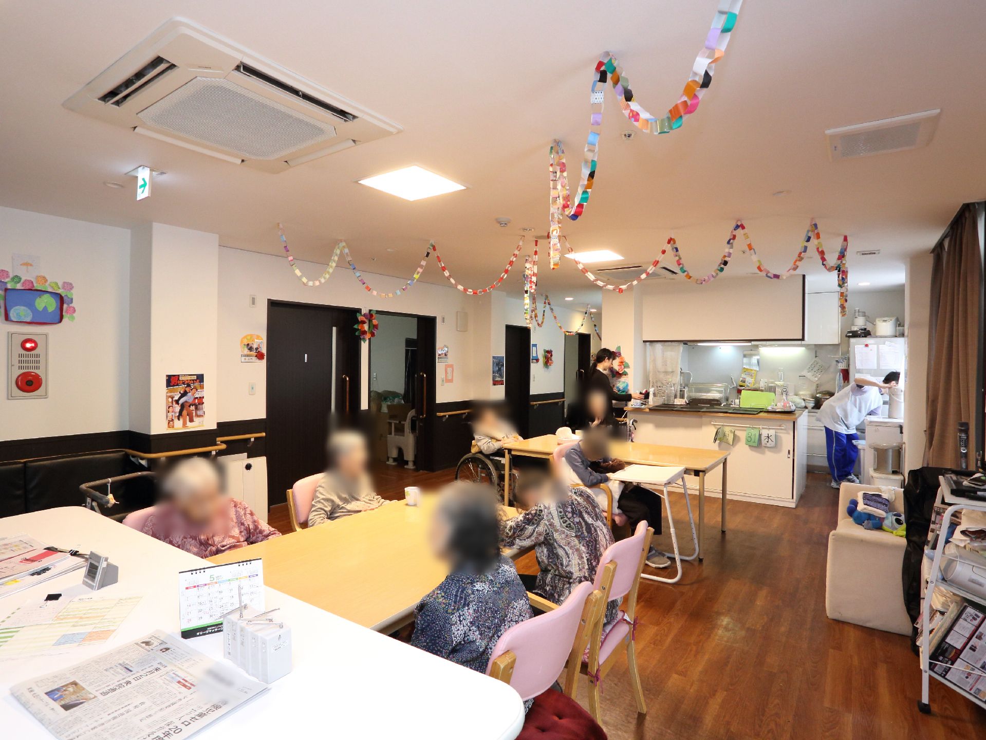 手前の棚には新聞がのっており奥の壁には非常ベルが設置してある。入居者達はテーブルを囲んでおり奥にはキッチンもある。