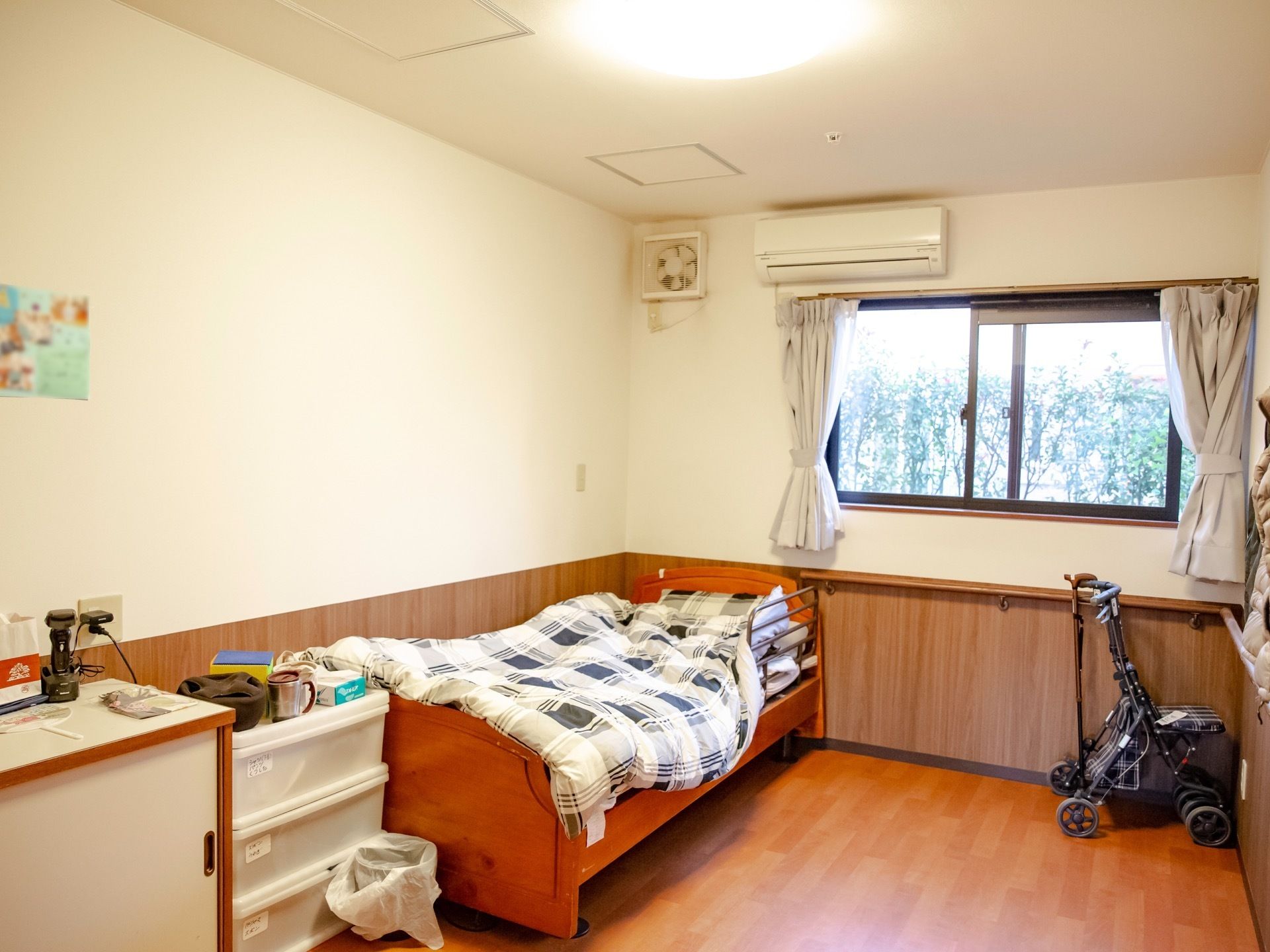 ベッドと棚が設置され、エアコンと換気扇がついている。窓からは外の木々が見える。また、歩行器と杖が部屋の隅に置かれている。