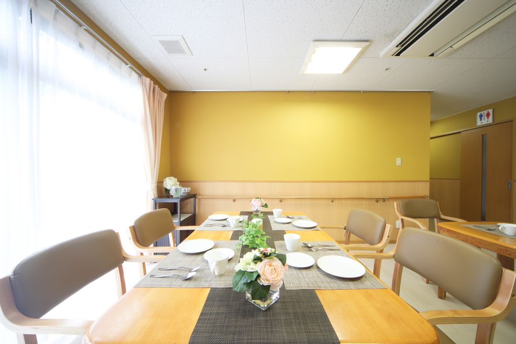 一面が窓の明るい部屋に、大きなダイニングテーブルがある。向かい合わせにピンク色の座面の椅子が合計８脚、並んでいる。壁に風景画が架かっている。