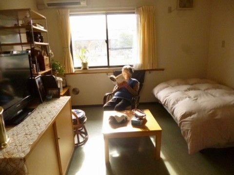 外光が入る窓際で、椅子に座った入居者様が本を読んでおられる。窓に向かって左側にはベッドがあり、反対側の壁には整理ダンスが置いてある。