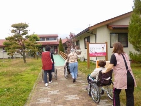 ３名の車椅子に乗った入居者様を、それぞれ一人ずつスタッフが介助して遊歩道を歩いている。周りには芝生と、庭木が見える。