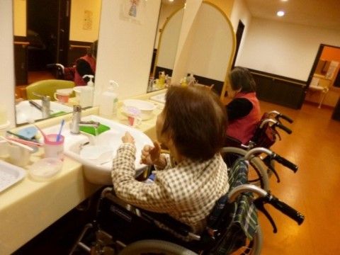 車椅子をご利用の方が２名並んで洗面台の前におられる。歯磨きや洗面などの身づくろいをなさっている。洗面台にはコップと歯磨きが置かれている。