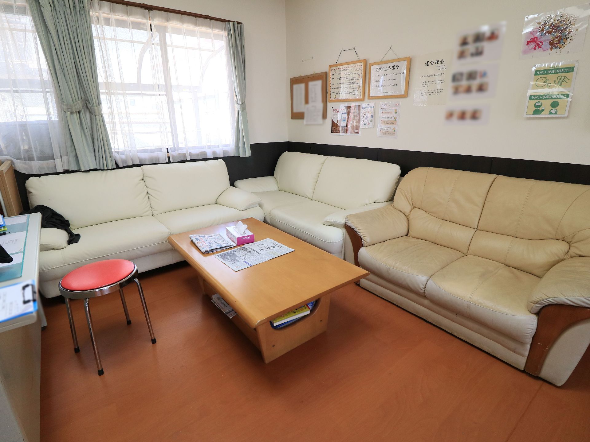 ソファとテーブルが置かれている。壁にはさまざまな掲示物が飾られている。窓にはカーテンが掛けられている。