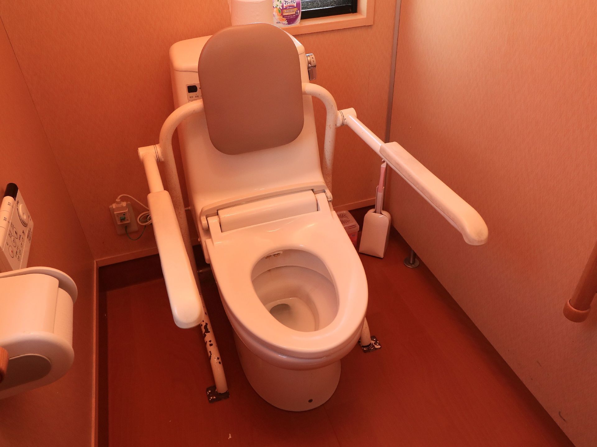 トイレの便座に蓋はなく、床に設置した器具で背もたれと手すりを設けている。温水洗浄機能の操作ボタンは壁に設置してある。